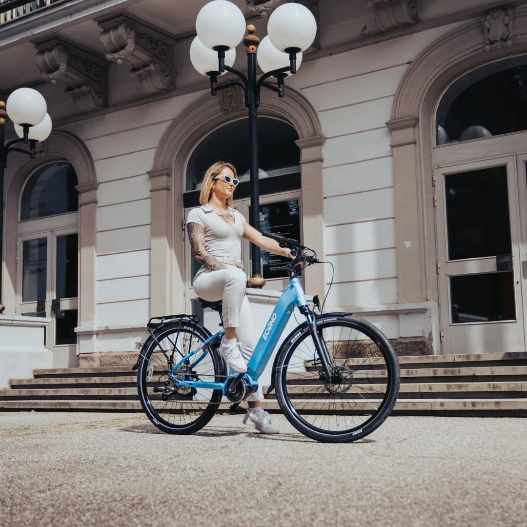 Na sliki je električno kolo znamke Econo, model Mare City. Kolo je v svetlo modri barvi in ima športen dizajn. Na kolesu sedi ženska, oblečena v svetlo obleko, ki se nahaja pred elegantno stavbo s stopnicami in svetilkami. V ozadju so vidne velike okenske odprtine, kar dodaja mestni ambient fotografiji.