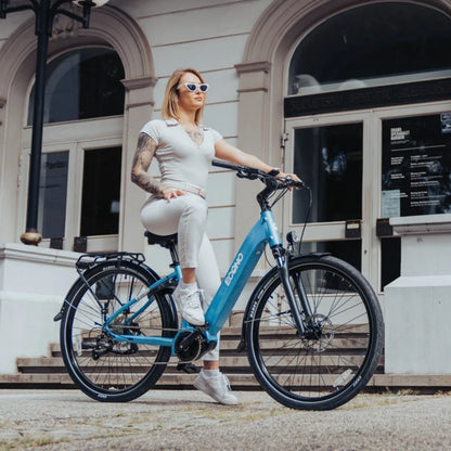 Na sliki je električno kolo znamke Econo, model Mare. Kolo je v svetlo modri barvi in ima sodoben dizajn. Na njem sedi mlada ženska, oblečena v svetle športne obleke, ter nosi sončna očala. V ozadju so vidne arhitekturne značilnosti stavbe, kar ustvarja prijetno urbano okolje.