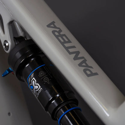 Izdelek je električno kolo, znamke Pantera, model Econo Pantera C8 140. Na sliki je vidna okvirna cev kolesa z logotipom Pantera ter del vzmetenja, ki vključuje amortizer s črno in modro barvo. Amortizer je opremljen z oznakami za nastavitev, kar nakazuje na njegovo funkcionalnost in prilagodljivost.