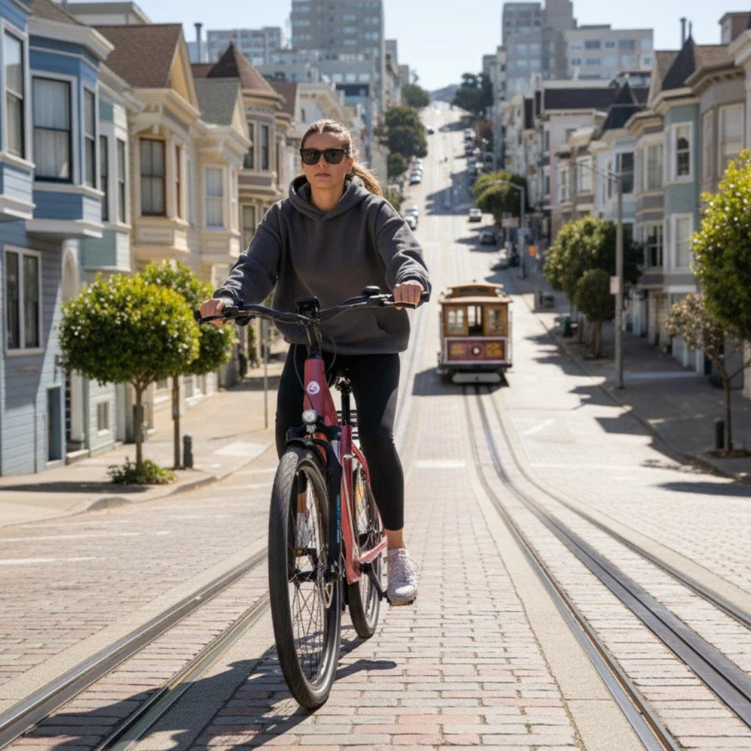 Izdelek je električno kolo, model Econo Apes EER4. Na sliki vidimo žensko, ki vozi električno kolo po strmi ulici, obdani z značilnimi hišami. V ozadju je vidna tramvajska proga in tramvaj, kar dodatno poudarja urbano okolje. Ženska nosi udobna oblačila in sončna očala, kar nakazuje na sproščen dan.