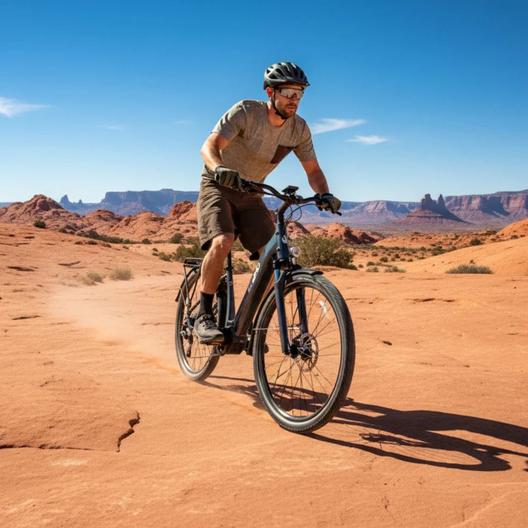 Na sliki je električno kolo, ki ga vozi moški v naravnem okolju. Kolo ima robustno zasnovo, primerno za vožnjo po terenu, in je opremljeno z dodatno opremo, kot je prtljažnik. Moški nosi čelado in zaščitna očala, kar kaže na aktivno in varno vožnjo. V ozadju so vidni skalnati hribi in modro nebo, kar ustvarja slikovito pokrajino.