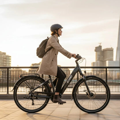 Na sliki je električno kolo znamke Econo, model Mare City. Kolo je črne barve in ima elegantno obliko, kar ga naredi primernega za mestno vožnjo. Na kolesu je vidna voznica, oblečena v svetlo plašč in s čelado, ki se vozi po pločniku ob reki, v ozadju pa so mestne zgradbe in sončni zahod. Kolo ima tudi prtljažnik, kar je praktično za prevoz stvari.