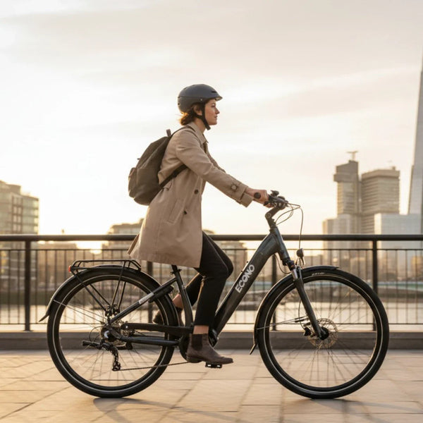Na sliki je električno kolo znamke Econo, model Mare City. Kolo je črne barve in ima elegantno obliko, kar ga naredi primernega za mestno vožnjo. Na kolesu je vidna voznica, oblečena v svetlo plašč in s čelado, ki se vozi po pločniku ob reki, v ozadju pa so mestne zgradbe in sončni zahod. Kolo ima tudi prtljažnik, kar je praktično za prevoz stvari.