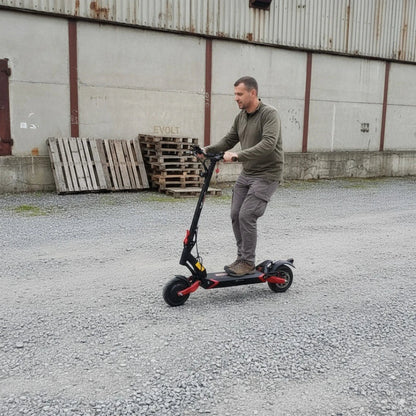 Na sliki je električni skiro, model Eco Speed 9X. Oseba na skiroju se vozi po makadamski površini v okolju z industrijskimi elementi, kot so lesene palete in stara zgradba v ozadju. Električni skiro ima črno in rdečo barvno kombinacijo ter široka kolesa, kar nakazuje na stabilnost in udobno vožnjo.