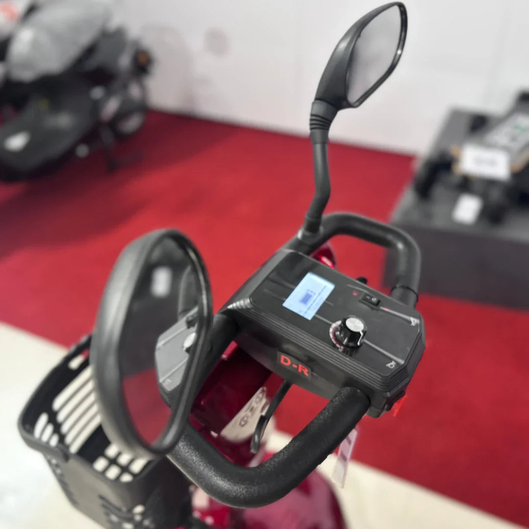 Izdelek na sliki je električni mobilni voziček, model Eco Speed Senior. Na sliki je vidna upravljalna enota z ročaji, ki vključuje zaslon in različne kontrolne gumbe. Poleg tega so na vozičku nameščeni tudi ogledali, kar omogoča boljšo vidljivost med vožnjo. Barva vozička je rdeča, kar daje izdelku privlačen videz.