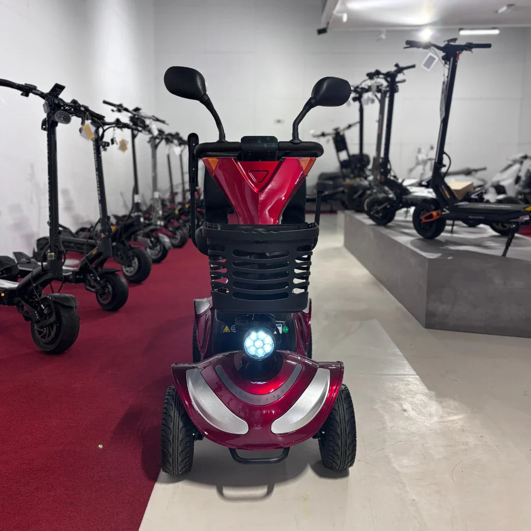 Izdelek na sliki je električni mobilni voziček, model Eco Speed Senior. Na sliki je vidna rdeča barva vozička, ki ima široke pnevmatike in svetlobno napravo spredaj. V ozadju so prikazani drugi električni skiroji, kar nakazuje, da gre za prodajno mesto ali razstavo. Voziček je zasnovan za udobno in varno vožnjo, kar je še posebej primerno za starejše uporabnike.