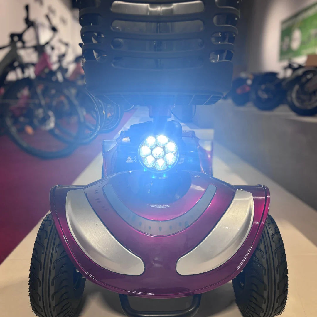 Izdelek je električni mobilni voziček, model Eco Speed Senior. Na sliki je vidna sprednja stran vozička z močnimi LED lučmi, ki osvetljujejo pot. Voziček ima široke gume, kar omogoča stabilno vožnjo, in je v privlačni vijolični barvi. V ozadju so vidni drugi kolesarski izdelki, kar nakazuje, da je slika posneta v trgovini ali razstavi.