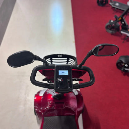 Izdelek na sliki je električni mobilni voziček, model Eco Speed Senior. Na sliki je prikazan nadzorni del vozička, kjer so vidni ročaji, dva ogledala ter zaslon, ki prikazuje informacije o vožnji. Barva vozička je rdeča, kar mu daje privlačen videz. V ozadju so vidni tudi drugi električni skiroji.