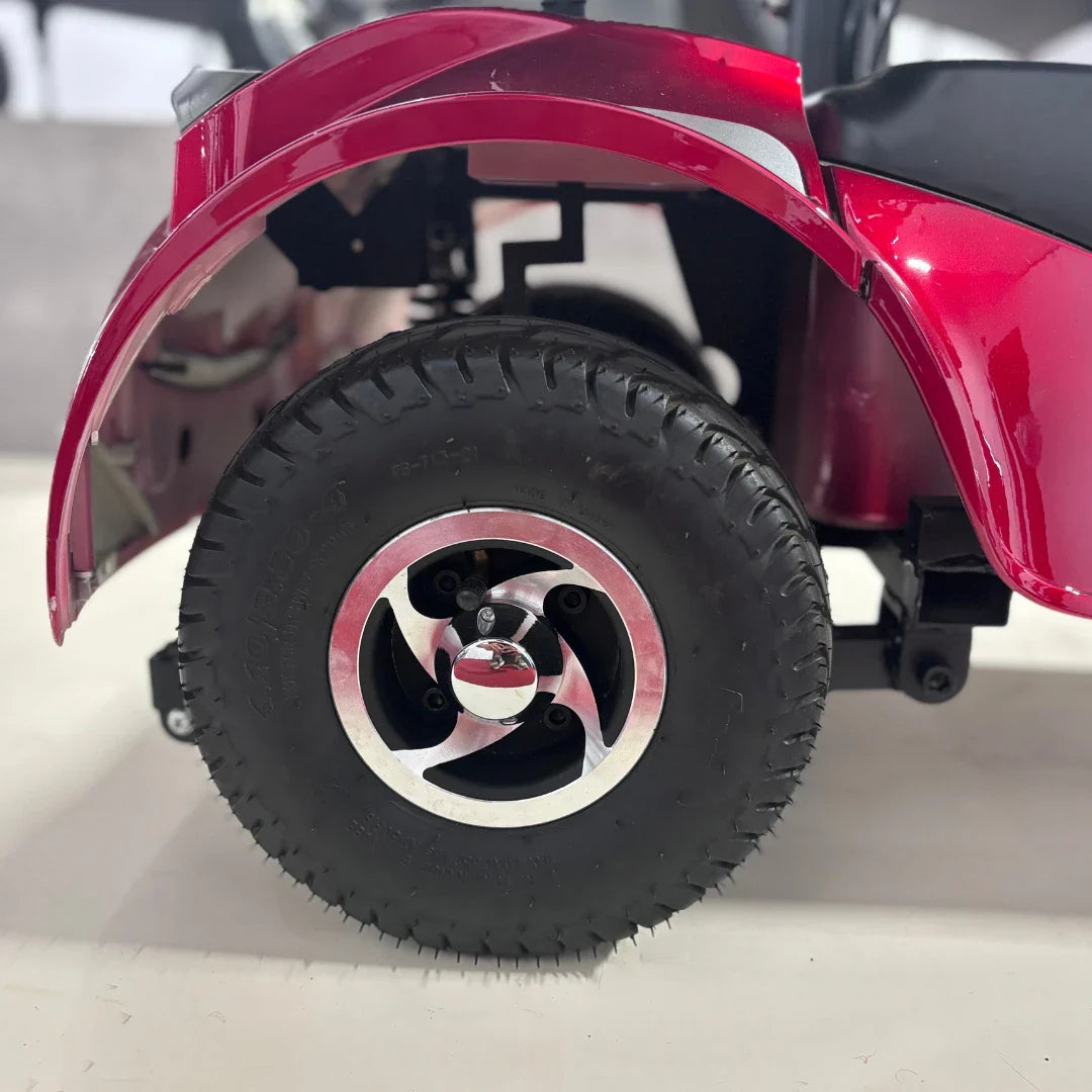 Izdelek je električni mobilni voziček, model Eco Speed Senior. Na sliki je vidna zadnja kolesa vozička, ki ima široko gumo in kromirano platišče. Barva vozička je rdeča, kar daje izdelku privlačen videz. Kolesa so zasnovana za stabilno vožnjo, kar je pomembno za udobje uporabnika.