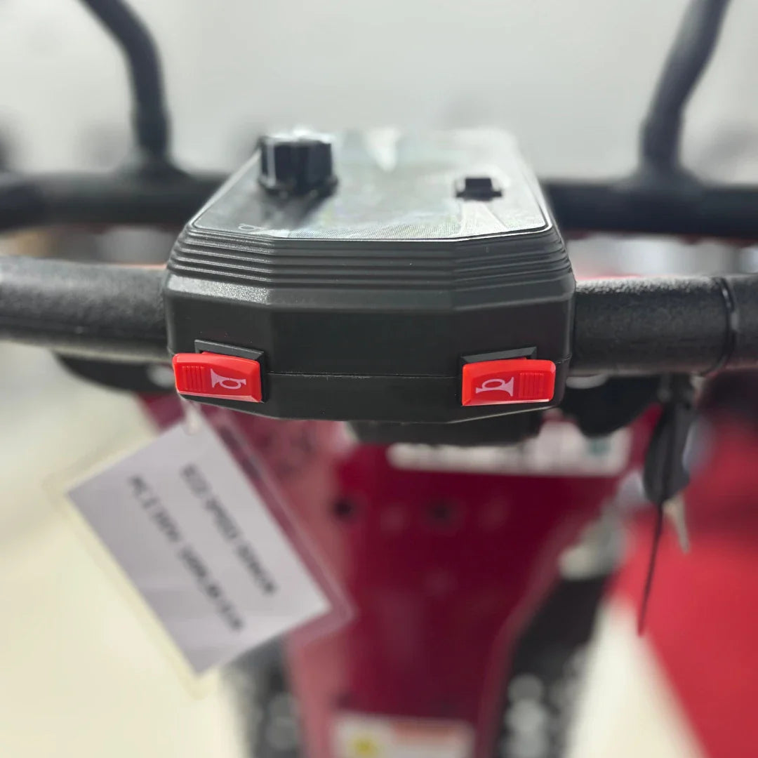 Izdelek je električni mobilni voziček, model Eco Speed Senior. Na sliki je prikazan del krmiljenja, ki vključuje gumbe za upravljanje in ključavnico. Vidna je tudi tekstura ročaja ter oznake na gumbih, kar nakazuje na enostavno uporabo. Barva vozička je rdeča, kar mu daje živahen videz.