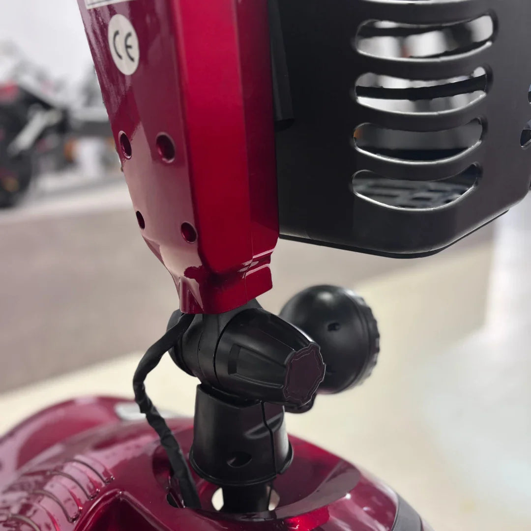 Izdelek je električni mobilni voziček, model Eco Speed Senior. Na sliki je prikazan del vozička, ki vključuje rdečo površino in črne komponente, kot so držalo in mehanizem za prilagajanje. Vidna je tudi oznaka CE, kar nakazuje, da izdelek izpolnjuje evropske varnostne standarde.