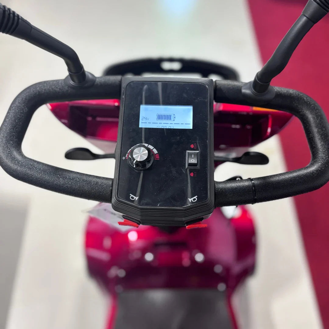 Izdelek je električni mobilni voziček, model Eco Speed Senior. Na sliki je vidna nadzorna plošča z digitalnim zaslonom, ki prikazuje nivo baterije, ter gumbi in nastavitvami za upravljanje. Voziček ima ergonomsko oblikovan krmilo, kar omogoča enostavno upravljanje in udobno vožnjo. Barva vozička je rdeča, kar mu daje privlačen videz.