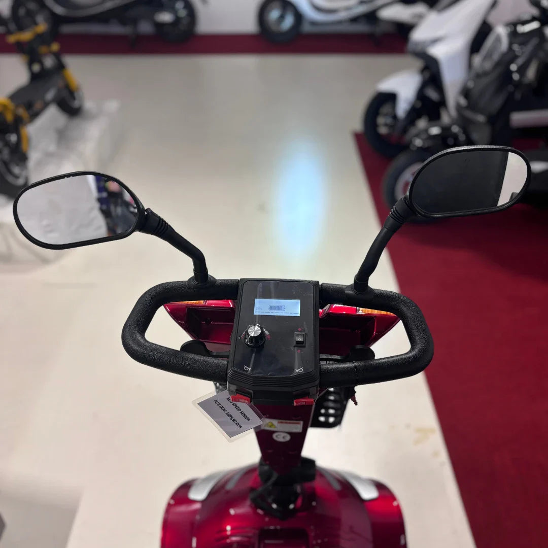 Izdelek je električni mobilni voziček, model Eco Speed Senior. Na sliki je vidna upravljalna plošča z zaslonom, ročaji in ogledali. Voziček je v rdeči barvi, kar mu daje privlačen videz. V ozadju so vidni tudi drugi električni vozili, kar nakazuje, da gre za prodajni prostor.