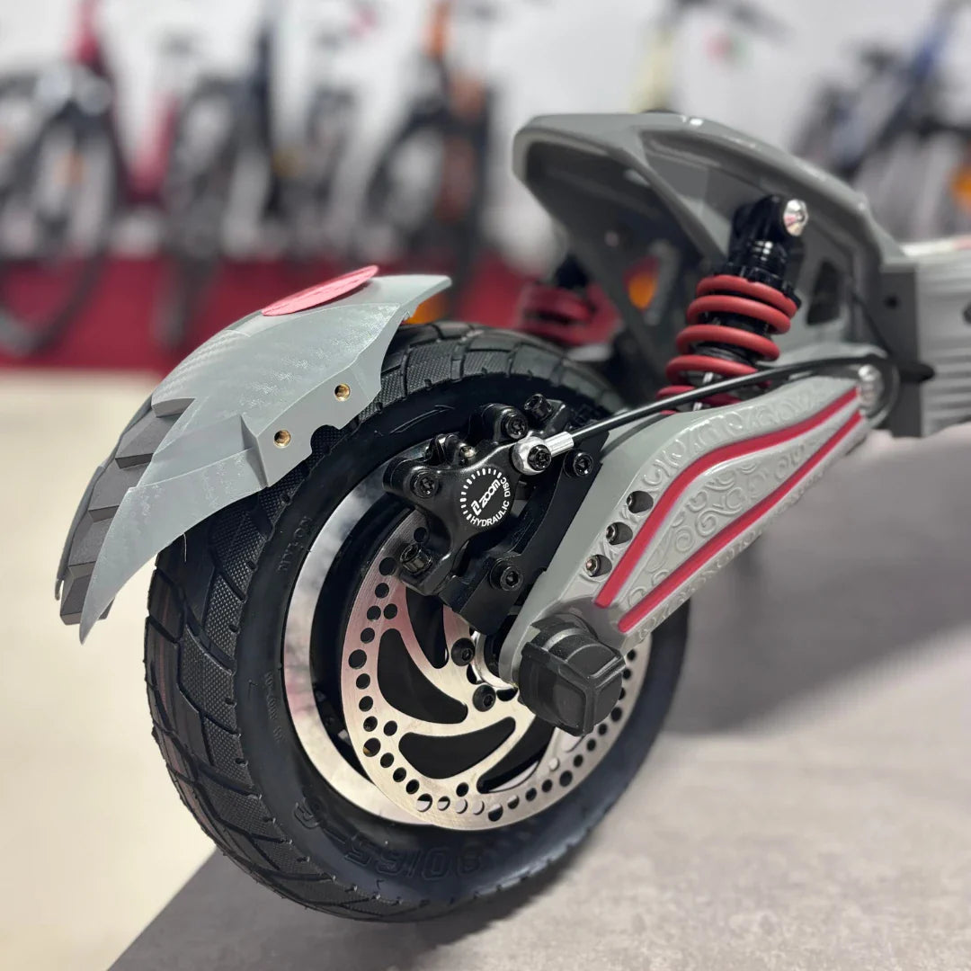 Izdelek na sliki je električni skiro, model Eco Speed 10X V2. Vidna je zadnja kolesa s širokim gumami, disk zavoro ter vzmetenje v rdeči barvi, kar nakazuje na dobro zmogljivost in udobje pri vožnji. Na ozadju so vidni tudi drugi kolesi, kar daje občutek o široki izbiri prevoznih sredstev v okolici.