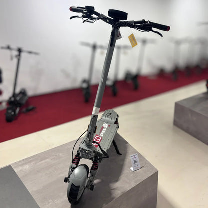 Izdelek na sliki je električni skiro, model Eco Speed 10X V2. Na skiroju so vidni široki sprednji in zadnji kolesi ter ročaji za upravljanje. V ozadju so vidni tudi drugi električni skiroji, kar nakazuje, da gre za razstavo ali prodajno mesto. Skira ima sodoben dizajn in je opremljen z amortizerji, kar povečuje udobje vožnje.