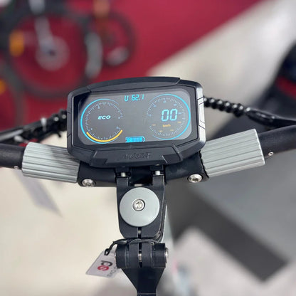 Izdelek je električni skiro, model Eco Speed 10X V2. Na sliki je prikazan nadzorni zaslon skiroja, ki prikazuje hitrost in način vožnje (ECO). Zaslon je opremljen z modrim ozadjem in analognimi merilniki, kar omogoča enostavno spremljanje podatkov med vožnjo. V ozadju so vidne tudi druge kolesarske naprave.