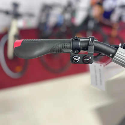 Izdelek je električni skiro, model Eco Speed 10X V2. Na sliki je vidna ročica skiroja s črnim in rdečim gumbom, ki verjetno služita za upravljanje funkcij, kot sta zvonec in morda način vožnje. V ozadju so vidni drugi kolesarski izdelki, kar nakazuje, da gre za razstavo ali prodajno mesto.