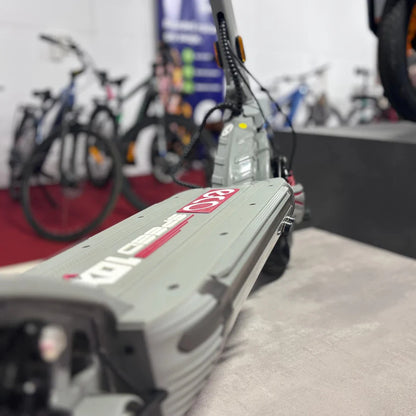 Na sliki je električni skiro model Eco Speed 10X V2. Vidna je ploščad skiroja z oznako "Eco Speed", ki je v sivi barvi. V ozadju so razporejeni drugi kolesarski izdelki, kar nakazuje, da je skiro del razstave ali prodajnega prostora. Skrbno oblikovan dizajn skiroja vključuje tudi detajle, kot so kabli in gumbi.