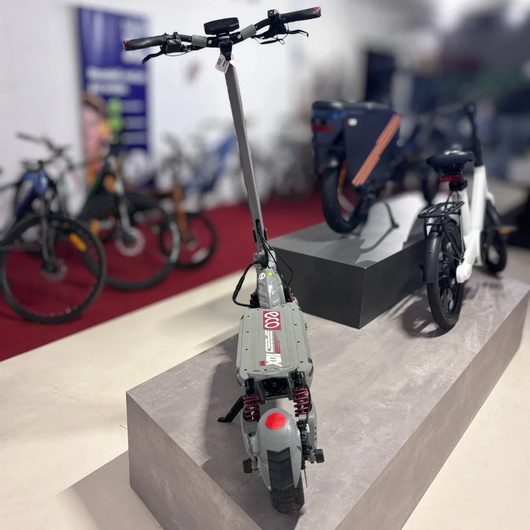 Na sliki je prikazan električni skiro model Eco Speed 10X V2. Izdelek ima robustno konstrukcijo, s širokimi pnevmatikami in vzmetenjem, kar nakazuje na udobno vožnjo. Na krmilu so vidni ročaji in nadzorna plošča, kar omogoča enostavno upravljanje. V ozadju so vidni tudi drugi kolesarski izdelki, kar daje občutek prodajnega prostora.