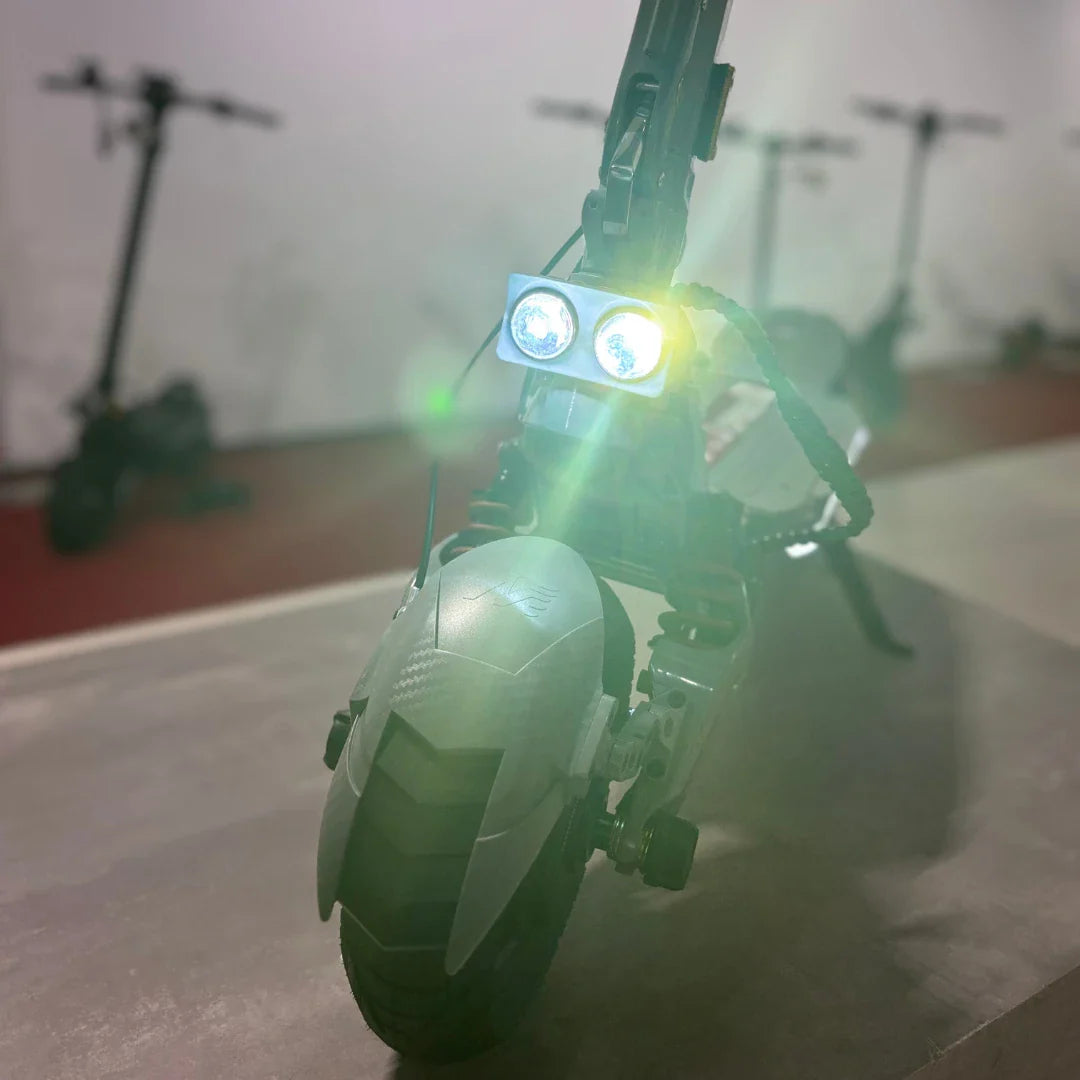 Izdelek je električni skiro, model Eco Speed 10X V2. Na sliki je prikazan sprednji del skiroja, ki ima dva svetlobna vira, kar nakazuje na možnost vožnje v temnejših pogojih. Vidna je tudi prednja guma in del okvirja, ki je zasnovan za stabilnost in udobje med vožnjo. V ozadju so vidni drugi električni skiroji.