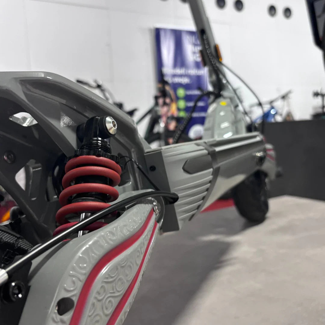 Na sliki je električni skiro, model Eco Speed 10X V2. Vidna je podrobnost vzmetenja s rdečimi vzmetmi ter del okvirja skiroja. Ozadje slike prikazuje druge električne naprave, kar nakazuje, da je skiro del razstave ali trgovine.