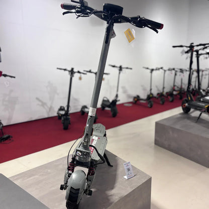 Izdelek na sliki je električni skiro, model Eco Speed 10X V2. Na sliki je vidna sprednja stran skiroja, ki ima robustno konstrukcijo in široke pnevmatike. Na krmilu so ročaji in nadzorna plošča, v ozadju pa so vidni tudi drugi električni skiroji, kar nakazuje, da gre za prodajno mesto. Barve skiroja so večinoma sivo in črno.