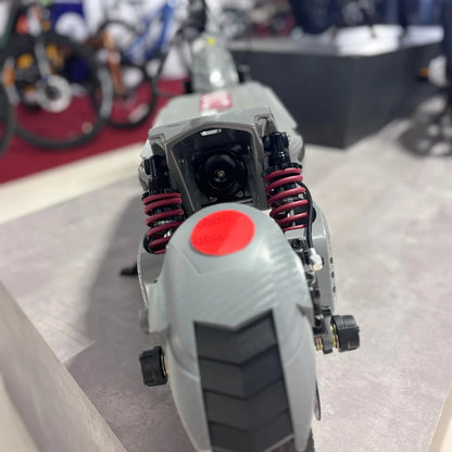 Izdelek je električni skiro, model Eco Speed 10X V2. Na sliki je vidna zadnja stran skiroja, kjer so prikazani amortizerji v rdeči barvi ter zadnje kolo s profilom za boljšo oprijemljivost. Oprema je v sivi barvi, kar daje sodoben videz. V ozadju so vidni tudi drugi kolesi, kar nakazuje, da je skiro postavljen na razstavi ali v trgovini.