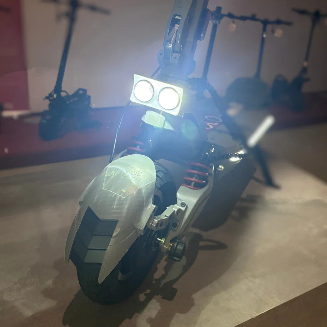 Na sliki je električni skiro model Eco Speed 10X V2. Vidna je sprednja stran skiroja, ki ima dva svetlobna vira, kar nakazuje na dobro osvetlitev za vožnjo v temi. Prav tako so opazne rdeče vzmeti, ki zagotavljajo boljšo stabilnost in udobje med vožnjo. Scooter ima robustno zasnovo in široko zadnjo pnevmatiko. V ozadju so vidni tudi drugi skiroji, kar nakazuje, da gre za razstavo ali prodajno mesto.