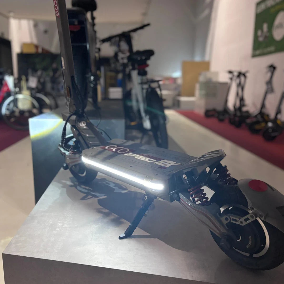 Izdelek na sliki je električni skiro, model Eco Speed 10X V2. Vidna je njegova robustna konstrukcija, s široko platformo in vzmetenjem, kar omogoča udobno vožnjo. Na sprednjem delu skiroja je nameščena LED luč, ki povečuje vidnost. V ozadju so vidni tudi drugi električni skiroji in kolesa, kar nakazuje, da gre za prodajno mesto ali razstavo.