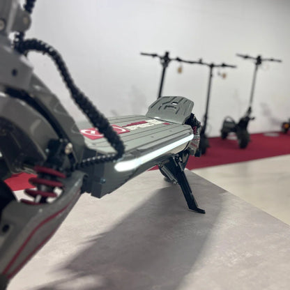 Na sliki je električni skiro model Eco Speed 10X V2. Vidna je platforma skiroja z značilno zasnovo in svetlobnim elementom, ki se nahaja ob robu. V ozadju so vidni še drugi električni skiroji, kar nakazuje, da gre za razstavo ali trgovinski prostor. Barve so pretežno sive s rdečimi detajli.