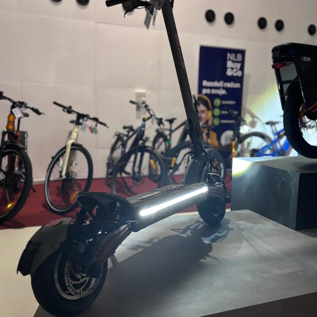 Na sliki je električni skiro, model Eco Speed 10X V2. Izdelek ima moderno zasnovo in je opremljen s svetlobnimi elementi, ki povečujejo vidnost. V ozadju so vidni tudi drugi kolesi, kar nakazuje, da je skiro del razstave ali trgovine s kolesi.