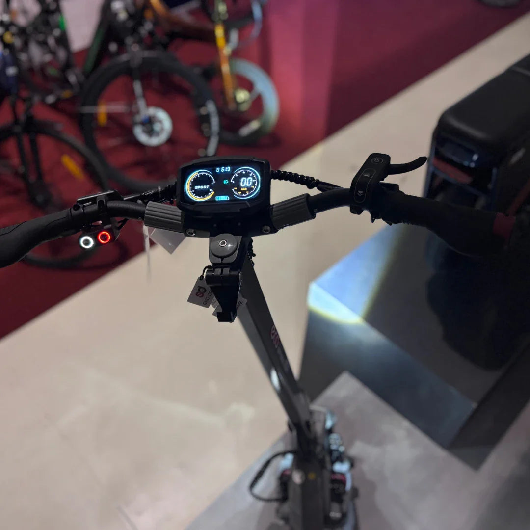 Izdelek je električni skiro, model Eco Speed 10X V2. Na sliki je prikazan nadzorni del skiroja z digitalnim zaslonom, ki prikazuje hitrost in način vožnje. Ročaji so opremljeni z gumboma za upravljanje, vidne so tudi luči. V ozadju so drugi kolesi, kar nakazuje, da je slika posneta v trgovini ali na razstavi.