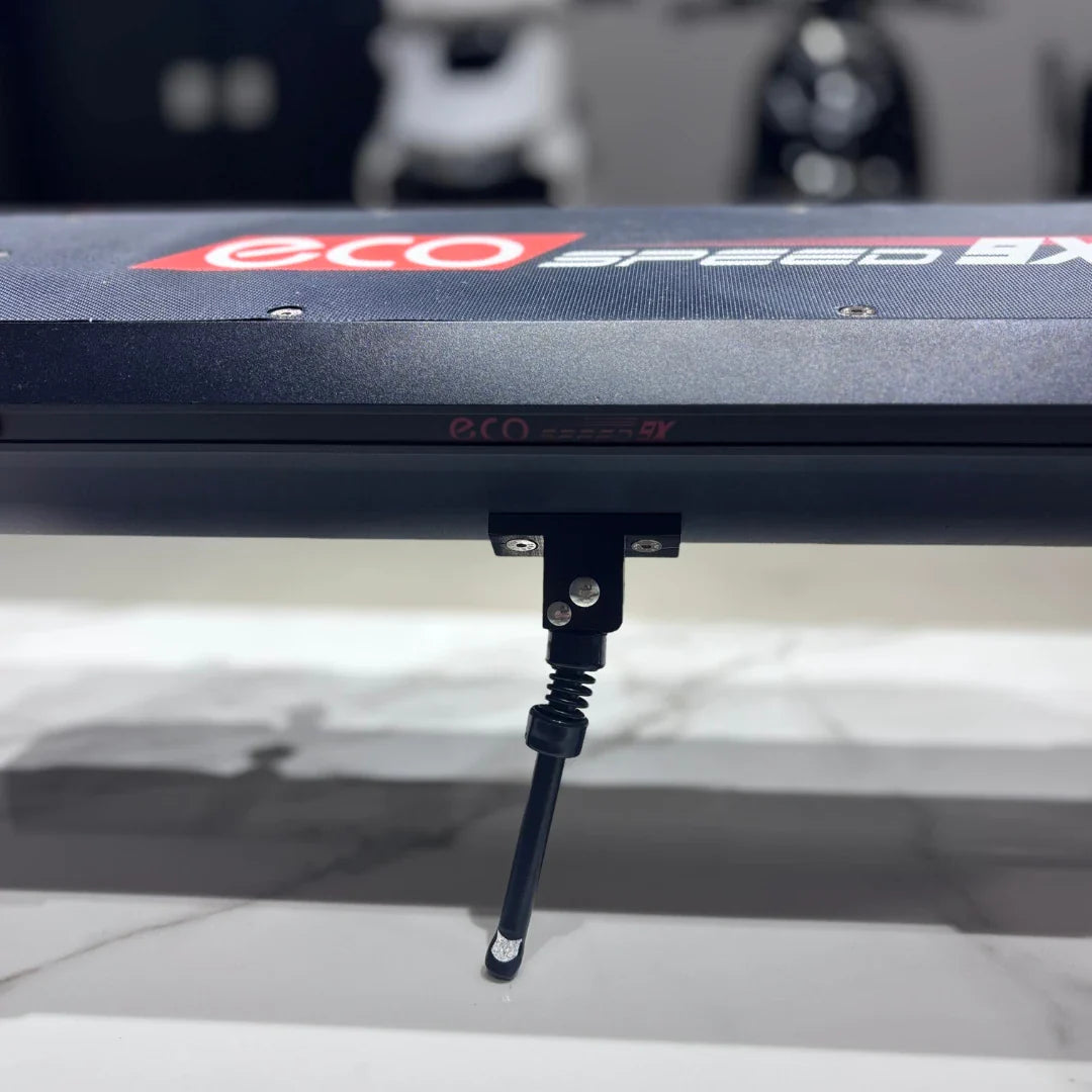 Izdelek na sliki je električni skiro, model Eco Speed 9X. Na sliki je vidna površina skiroja z logotipom "eco" in "speed 9X", ter del mehanizma, ki je verjetno povezan s sistemom za zlaganje ali stabilizacijo. Ozadje je zamegljeno, kar nakazuje, da je izdelek postavljen v trgovinskem okolju.