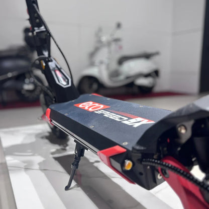 Izdelek je električni skiro, model Eco Speed 9X. Na sliki je vidna platforma skiroja z značilnim logotipom in barvno kombinacijo črne in rdeče. Opremljen je z nosilcem za stojnico, kar nakazuje na funkcionalnost in stabilnost. V ozadju so vidni tudi drugi električni prevozni sredstvi.