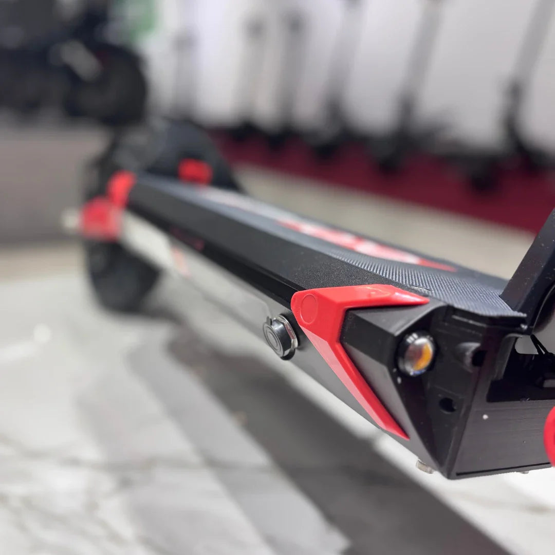 Na sliki je električni skiro model Eco Speed 9X. Vidna je ploščad skiroja, ki je opremljena z rdečimi detajli in gumirano površino za boljši oprijem. V ozadju so vidni tudi drugi električni skiroji, kar nakazuje, da gre za razstavni prostor. Celoten dizajn je športen in sodoben.