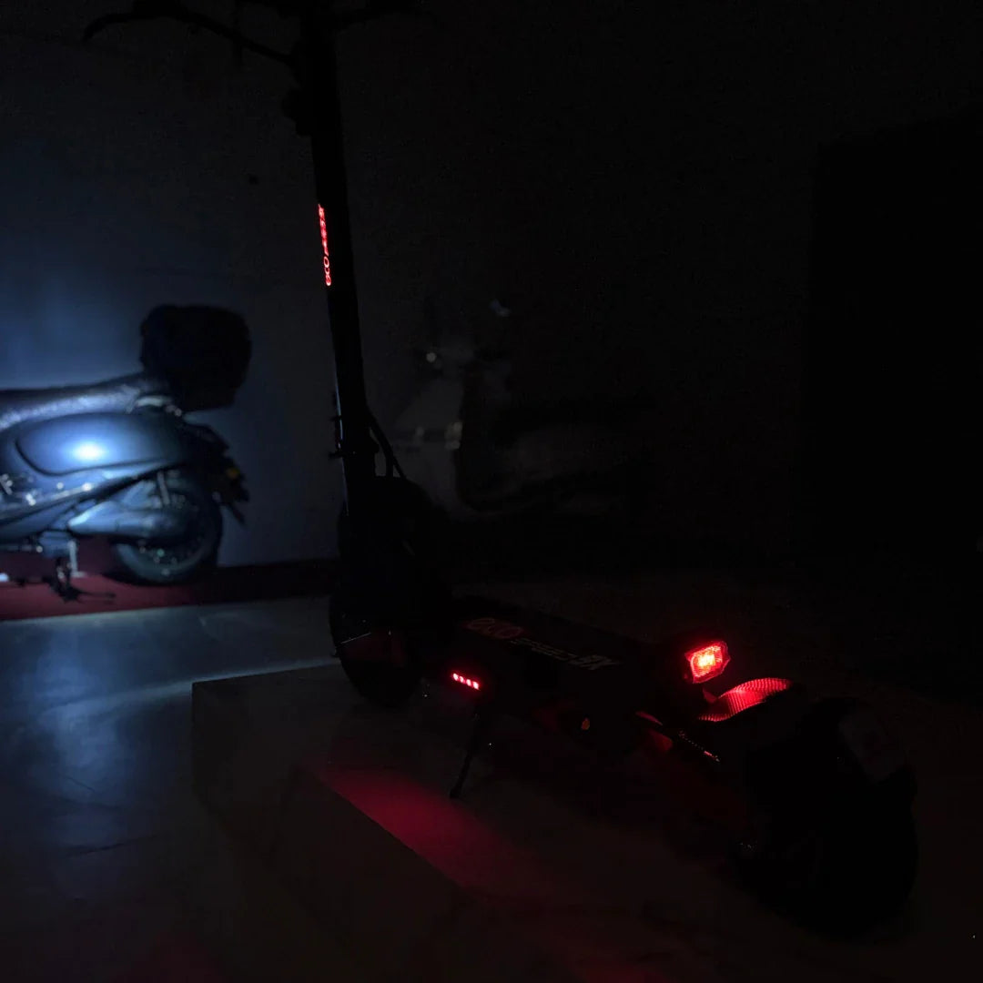 Na sliki je električni skiro model Eco Speed 9X. Vidna je zadnja stran skiroja, ki ima rdeče luči za boljšo vidnost v temi. V ozadju se vidi tudi motorno kolo, kar daje dodatno dimenzijo prostoru. Skupna osvetlitev okolice je temna, kar poudarja detajle skiroja.