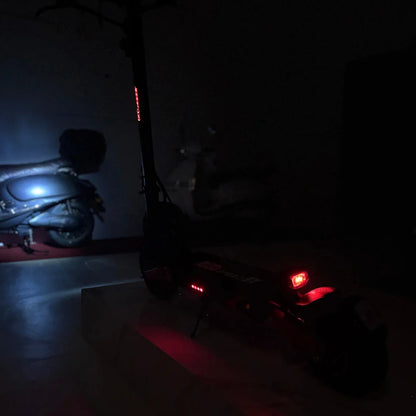 Na sliki je električni skiro model Eco Speed 9X. Vidna je zadnja stran skiroja, ki ima rdeče luči za boljšo vidnost v temi. V ozadju se vidi tudi motorno kolo, kar daje dodatno dimenzijo prostoru. Skupna osvetlitev okolice je temna, kar poudarja detajle skiroja.