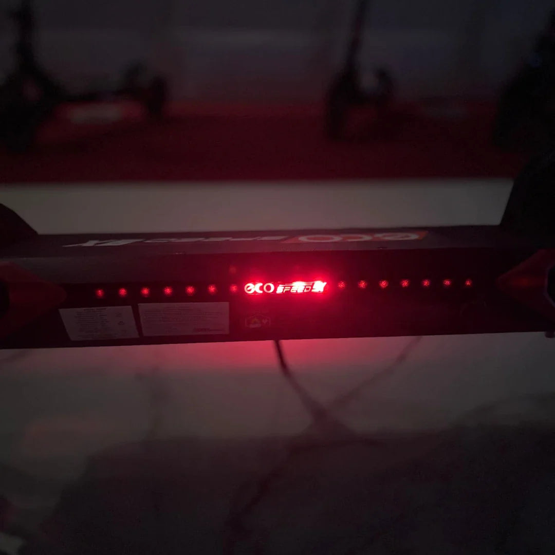 Izdelek na sliki je električni skiro, model Eco Speed 9X. Na sliki je vidna zgornja plošča skiroja, ki ima rdeče LED luči ter oznako znamke. V ozadju so vidni tudi drugi električni skiroji. Plošča vsebuje tudi informacije o delovanju in specifikacijah skiroja.