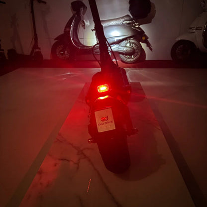 Na sliki je električni skiro model "Eko Drive". Vidna je zadnja stran skiroja, kjer se nahaja rdeča lučka, kar nakazuje na njegovo funkcionalnost in varnost pri vožnji v temi. V ozadju so še drugi skiroji in motorna kolesa, kar daje občutek urbanega okolja.