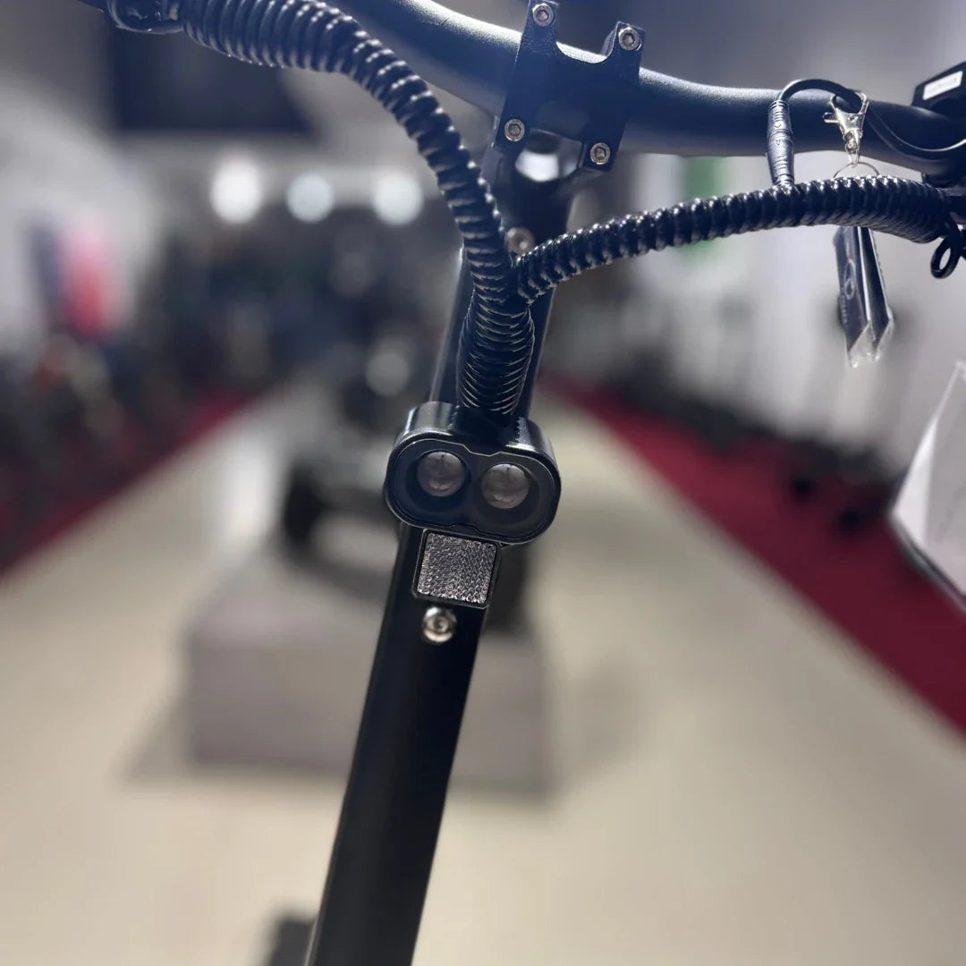 Izdelek na sliki je električni skiro, model Eco Speed 9X. Na sliki je prikazan del krmila, ki vključuje luči in odsevnik. Ozadje je nekoliko zamegljeno, kar nakazuje, da je skiro postavljen v trgovini, kjer so vidni tudi drugi električni skiroji. Skupna zasnova je moderna in funkcionalna.