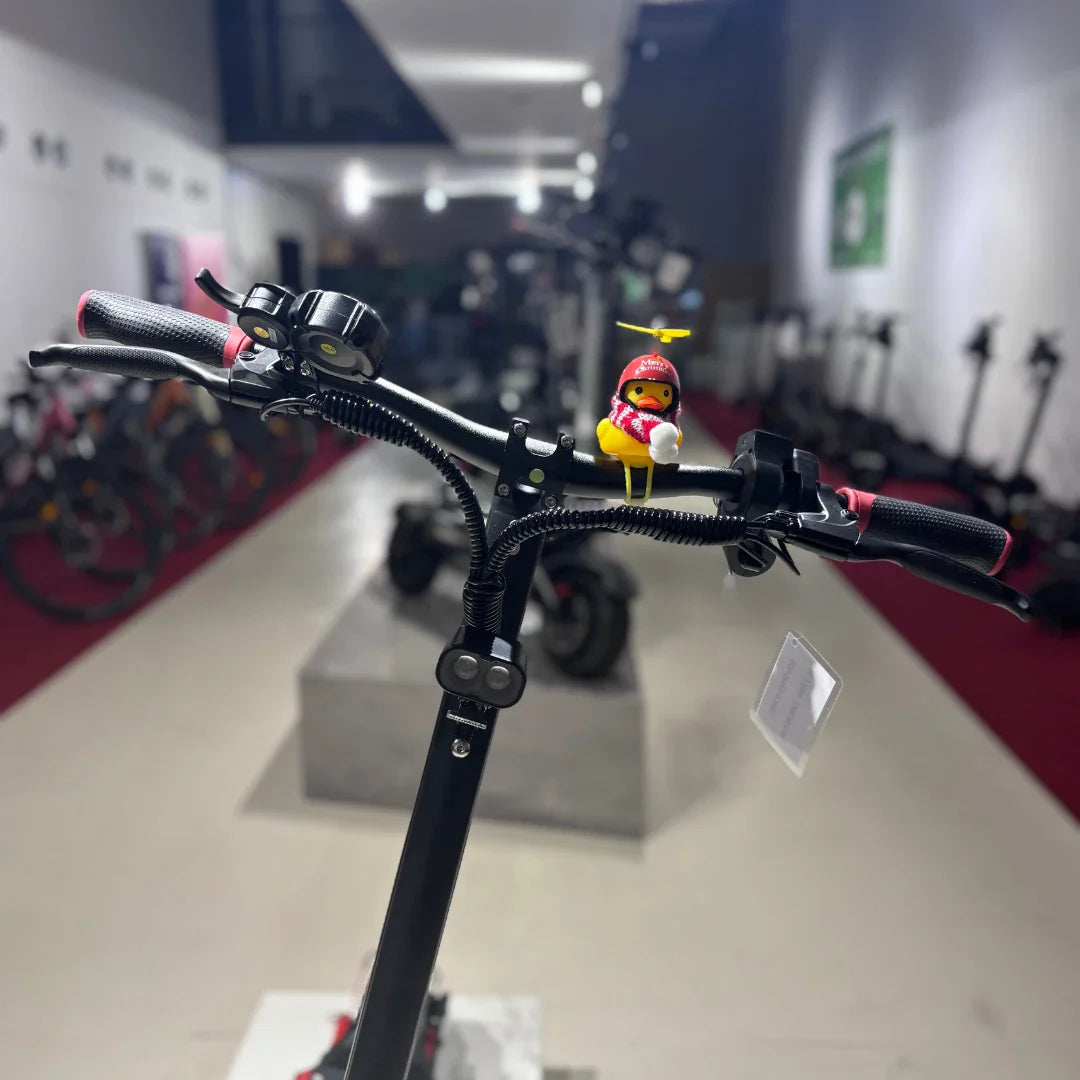 Na sliki je električni skiro model "Eco Speed 9". Vidni so krmili s črnimi ročaji in dodatkom v obliki majhne igrače, ki je postavljena na vrhu. V ozadju so vidni drugi električni skiroji, kar nakazuje, da gre za prodajno mesto ali razstavo. Skuter ima sodoben dizajn in je pripravljen za uporabo.