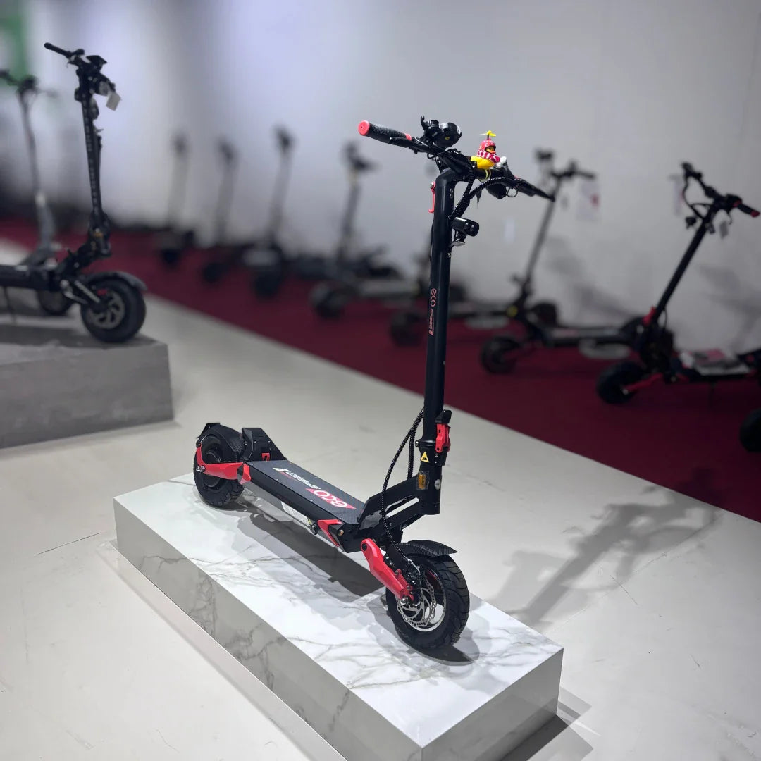 Na sliki je električni skiro model Eco Speed 9. Izdelek je postavljen na podstavek, kar omogoča boljši pregled nad njegovim dizajnom. Skiro ima črno in rdečo barvno kombinacijo ter široka kolesa, kar nakazuje na stabilnost in udobno vožnjo. V ozadju so vidni tudi drugi električni skiroji, kar kaže na raznoliko izbiro v trgovini.