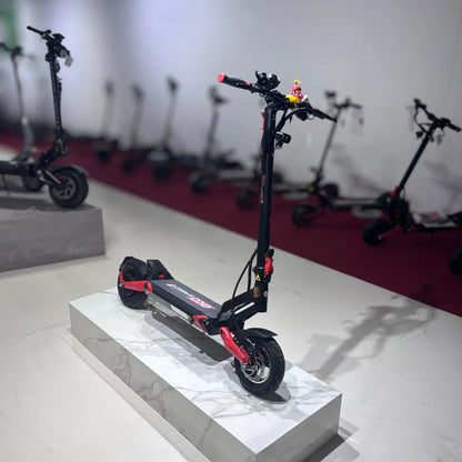 Na sliki je električni skiro model Eco Speed 9. Izdelek je postavljen na podstavek, kar omogoča boljši pregled nad njegovim dizajnom. Skiro ima črno in rdečo barvno kombinacijo ter široka kolesa, kar nakazuje na stabilnost in udobno vožnjo. V ozadju so vidni tudi drugi električni skiroji, kar kaže na raznoliko izbiro v trgovini.