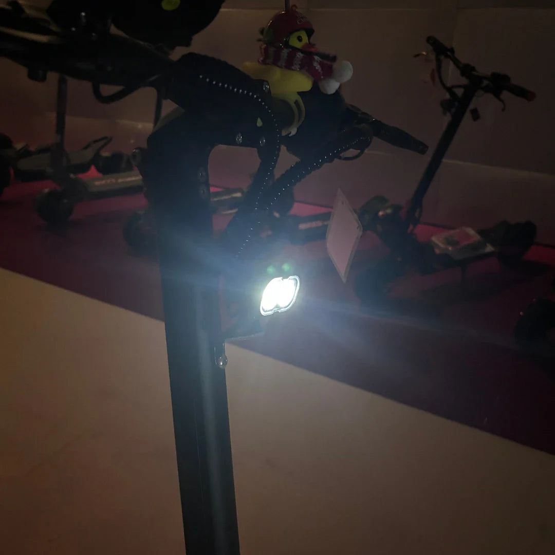 Na sliki je prikazan električni skiro model "Eco Speed 9". Na krmilu skiroja je vidna lučka, ki osvetljuje pot, kar je pomembno za varno vožnjo v temi. Na vrhu krmila je tudi plišasta igrača, kar dodaja osebno noto in barvitost. V ozadju so vidni še drugi električni skiroji.