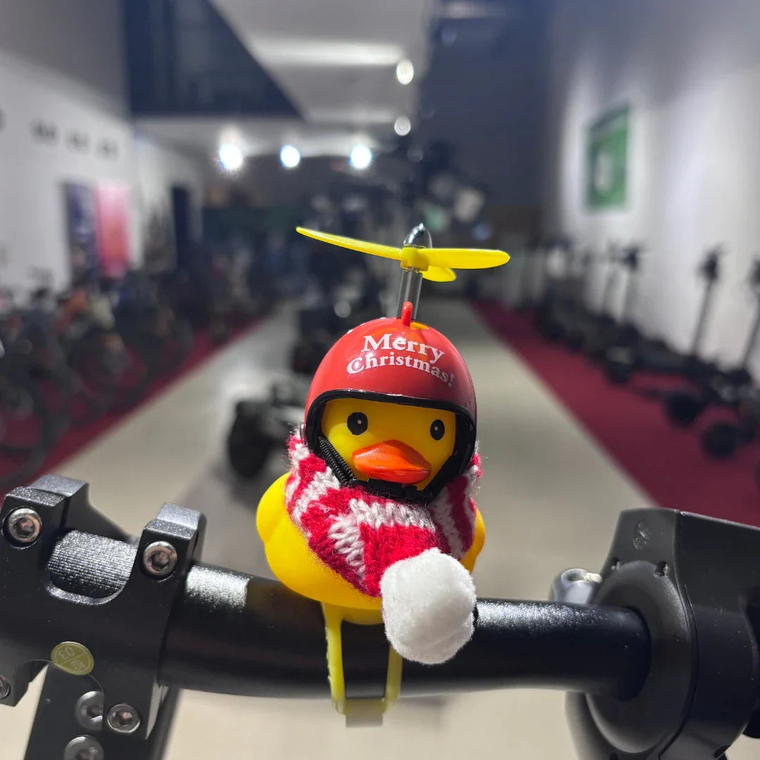 Na sliki je električni skiro, model Eco Speed 9. Na krmilu skiroja je prikazana igračka v obliki rumene račke, ki nosi božična oblačila, vključno s čelado in šalom. V ozadju so vidni drugi električni skiroji, kar nakazuje, da je slika posneta v trgovini ali razstavi.