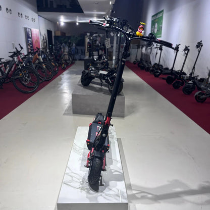 Na sliki je električni skiro model Eco Speed 9. Izdelek je postavljen na belem podstavku v trgovini, v ozadju pa so vidni drugi električni skiroji in kolesa. Scoot je črne barve z rdečimi detajli, na krmilu pa je pritrjen majhen okras. Skiro ima široke pnevmatike, kar nakazuje na dobro stabilnost in udobje pri vožnji.