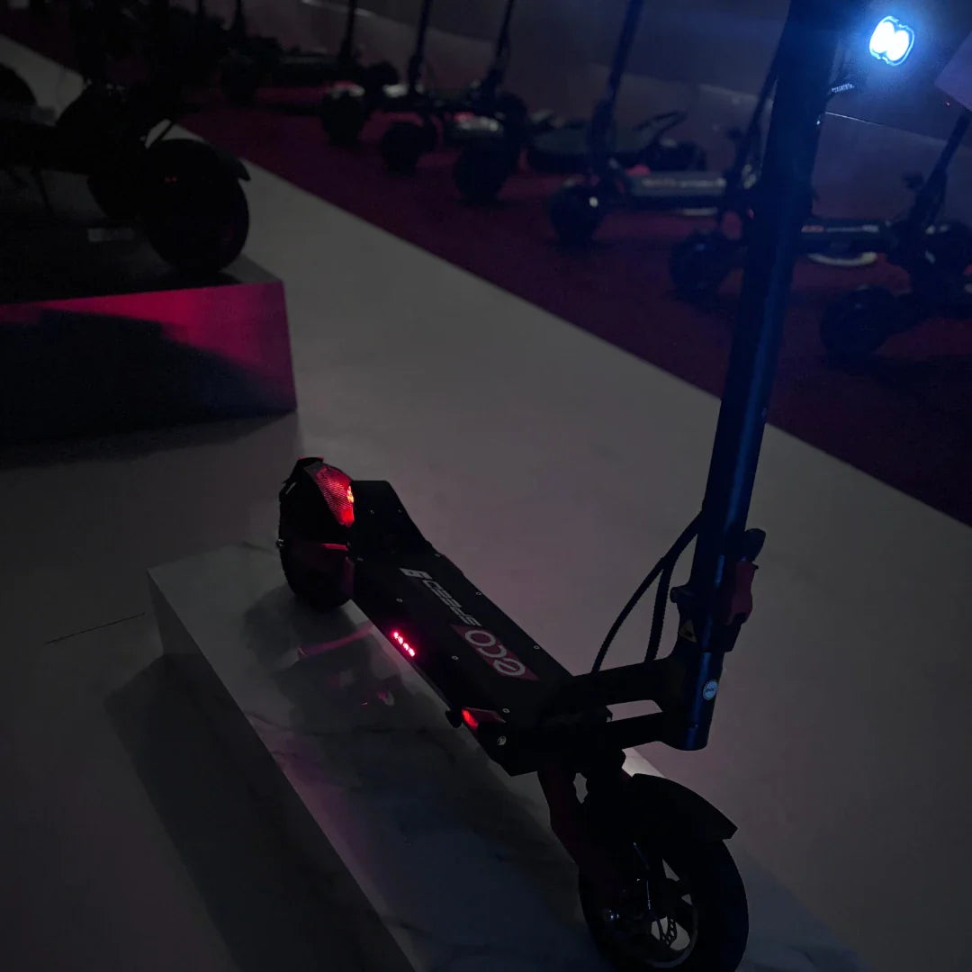 Na sliki je električni skiro model Eco Speed 9. Izdelek je črne barve in je postavljen na svetli podlagi, kar omogoča, da izstopajo detajli, kot so rdeče in modre luči. V ozadju so vidni tudi drugi električni skiroji, kar nakazuje, da gre za razstavo ali prodajno mesto. Scooter ima sodoben dizajn in je opremljen z LED lučmi, kar povečuje njegovo vidnost v temi.