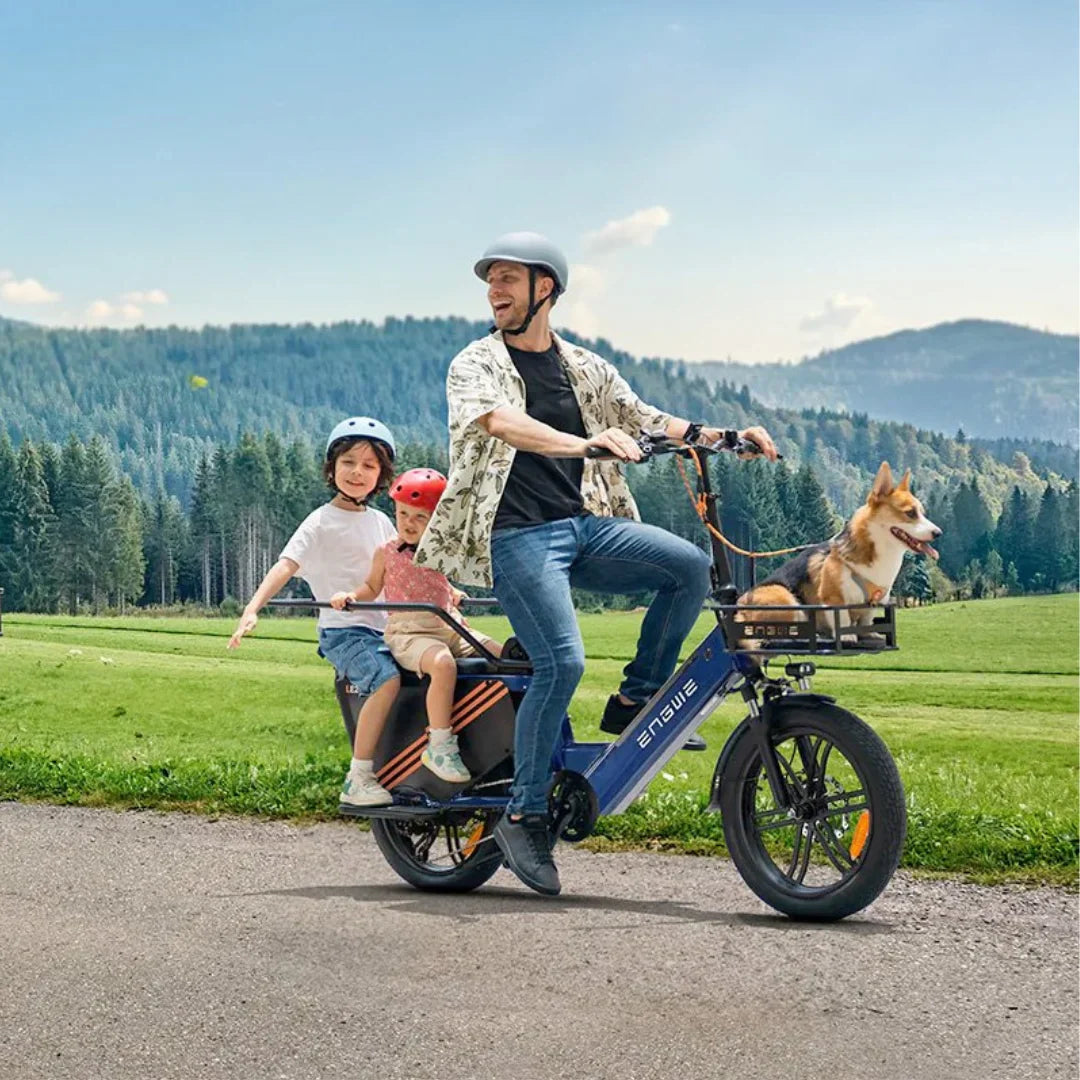 Izdelek na sliki je električno kolo, model Engwe LE20. Na sliki je moški, ki vozi kolo po podeželski poti, medtem ko prevozi dva otroka, enega spredaj in drugega zadaj. Na zadnjem delu kolesa je tudi košara, v kateri sedi pes. Okoliška narava je zelena in slikovita, kar ustvarja prijetno vzdušje za vožnjo.