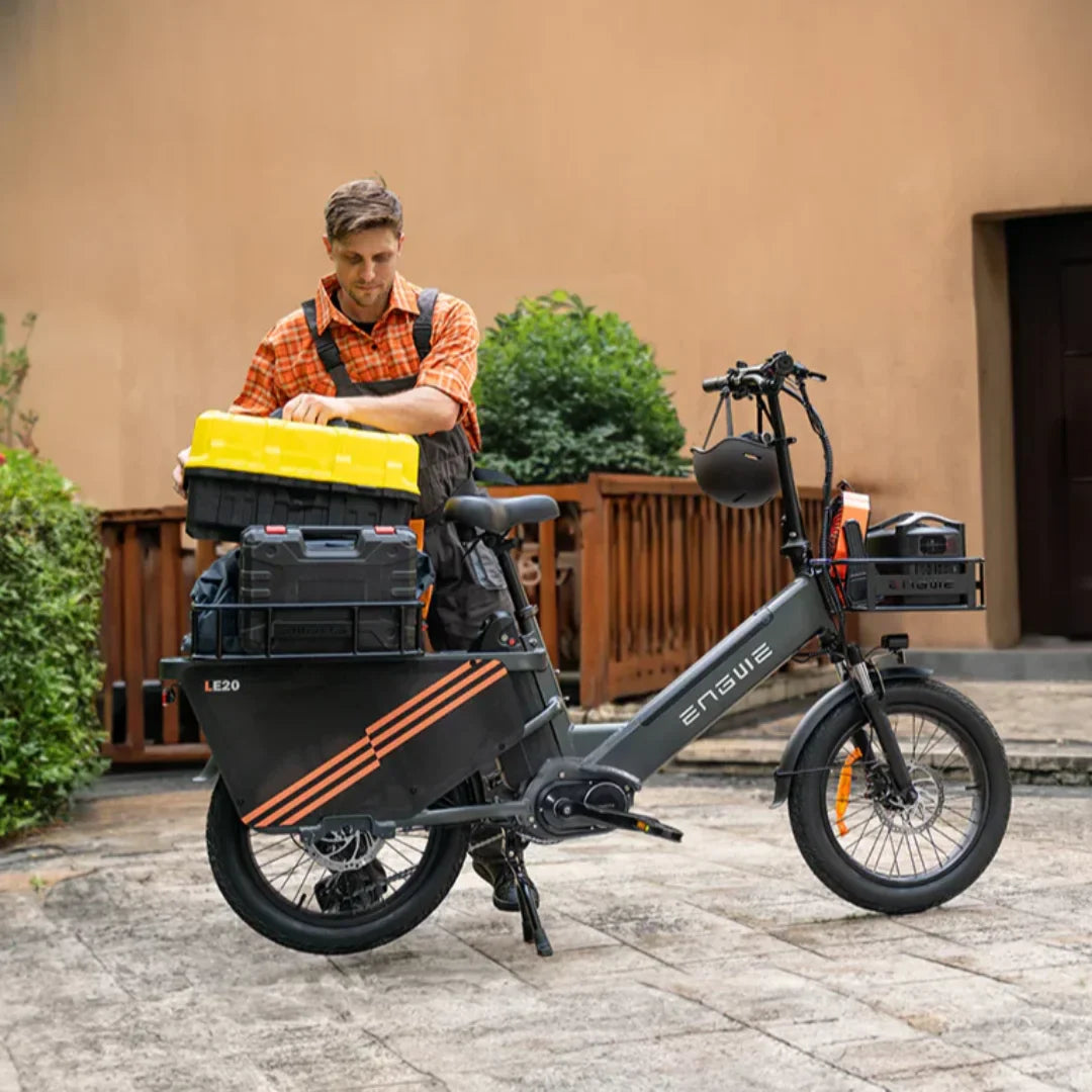 Na sliki je električno kolo znamke Engwe, model LE20. Kolo je opremljeno z različnimi škatlami za shranjevanje, kar nakazuje na njegovo praktično uporabo. Oseba na sliki se ukvarja z organizacijo opreme, kar kaže na uporabnost kolesa za prevoz orodja ali drugih predmetov. Kolo ima robustno zasnovo in je primerno za mestno vožnjo ali dostavo.