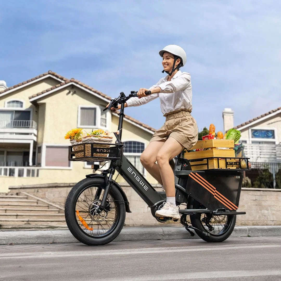 Na sliki je električno kolo znamke Engwe, model LE20. Kolo je opremljeno z dvema košarama, v katerih so vidni različni živilski izdelki, kar nakazuje, da je primerno za prevoz nakupov. Oseba, ki vozi kolo, nosi čelado in se nahaja na cesti pred stanovanjskimi hišami.